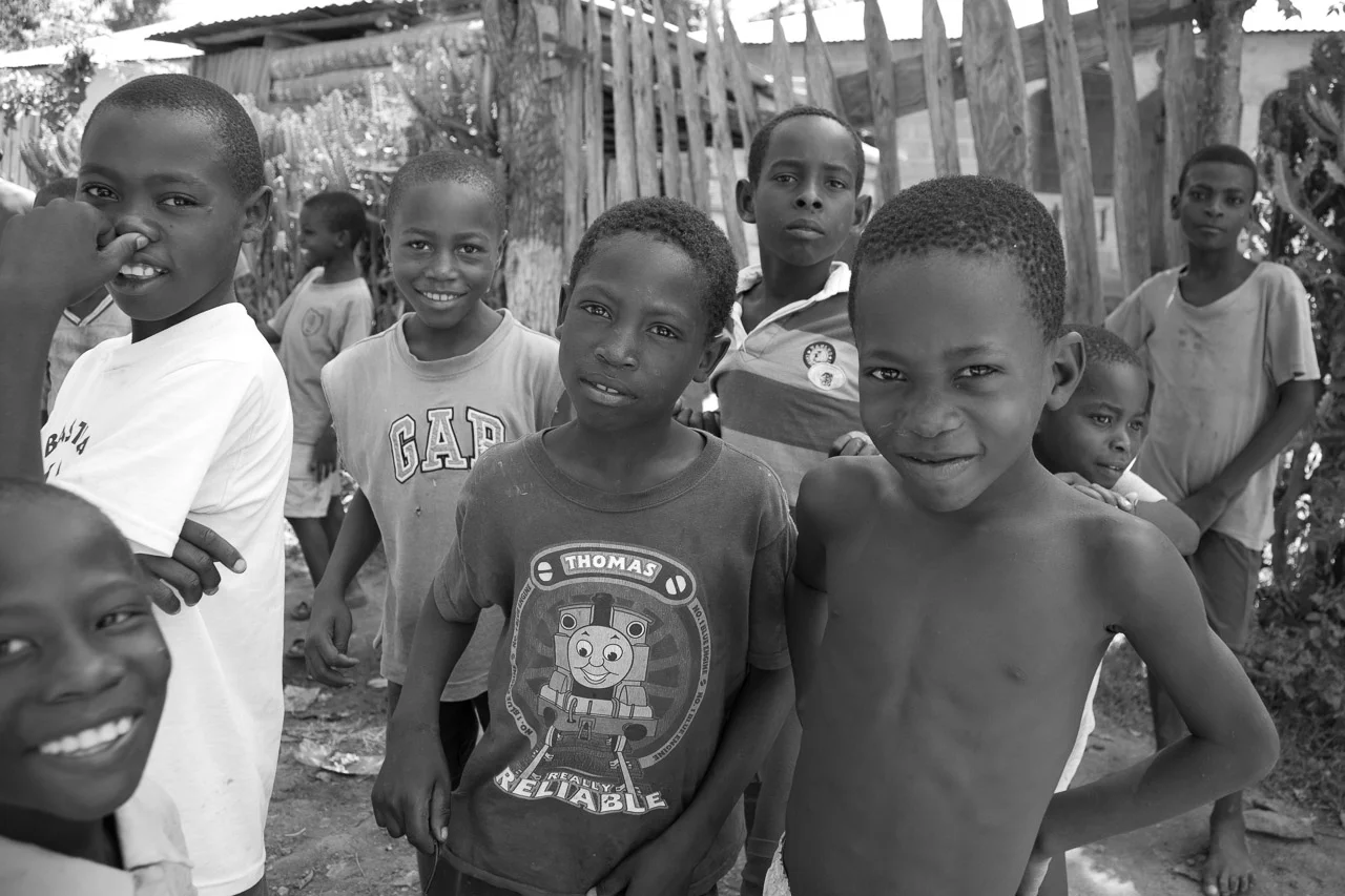 Group of African children standing outdoors, smiling and looking at the camera, with makeshift wooden structures in the background.
