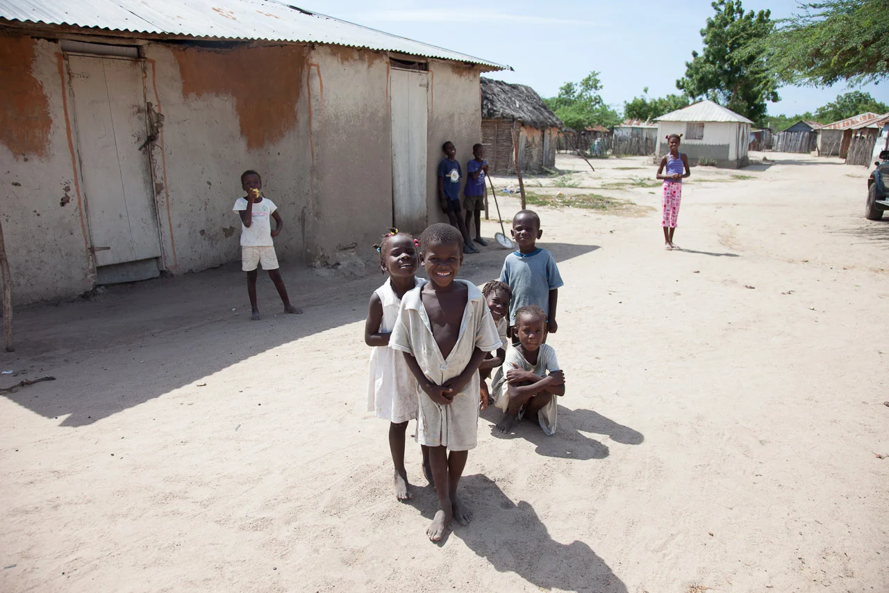 Six children are outside in a rural area with dirt ground, surrounded by simple buildings and trees. Four children are standing in the foreground, smiling and facing the camera. Two children are in the background near an old building, and one girl st