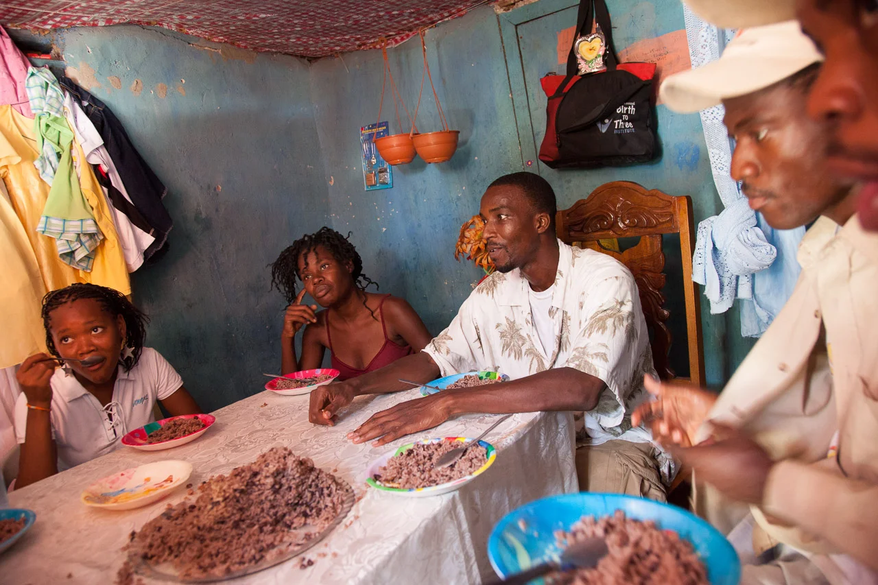 Family gathered around a dining table with a large pile of cooked rice, eating and talking in a modest room with blue walls and hanging clothes.
