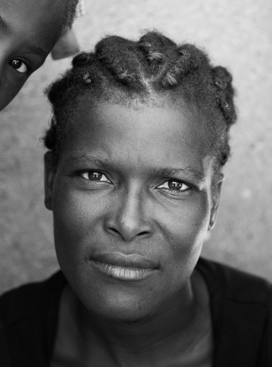 Close-up of a woman with braided hair, looking directly at the camera, with part of a child's face visible in the top left corner.
