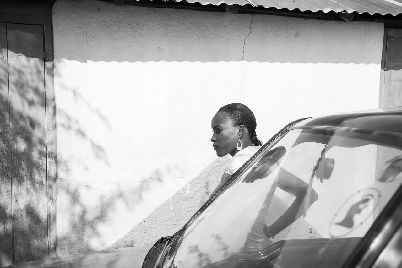 Side profile of a woman with earrings leaning on a car with a reflective windshield, against a textured wall with a door, in black and white.