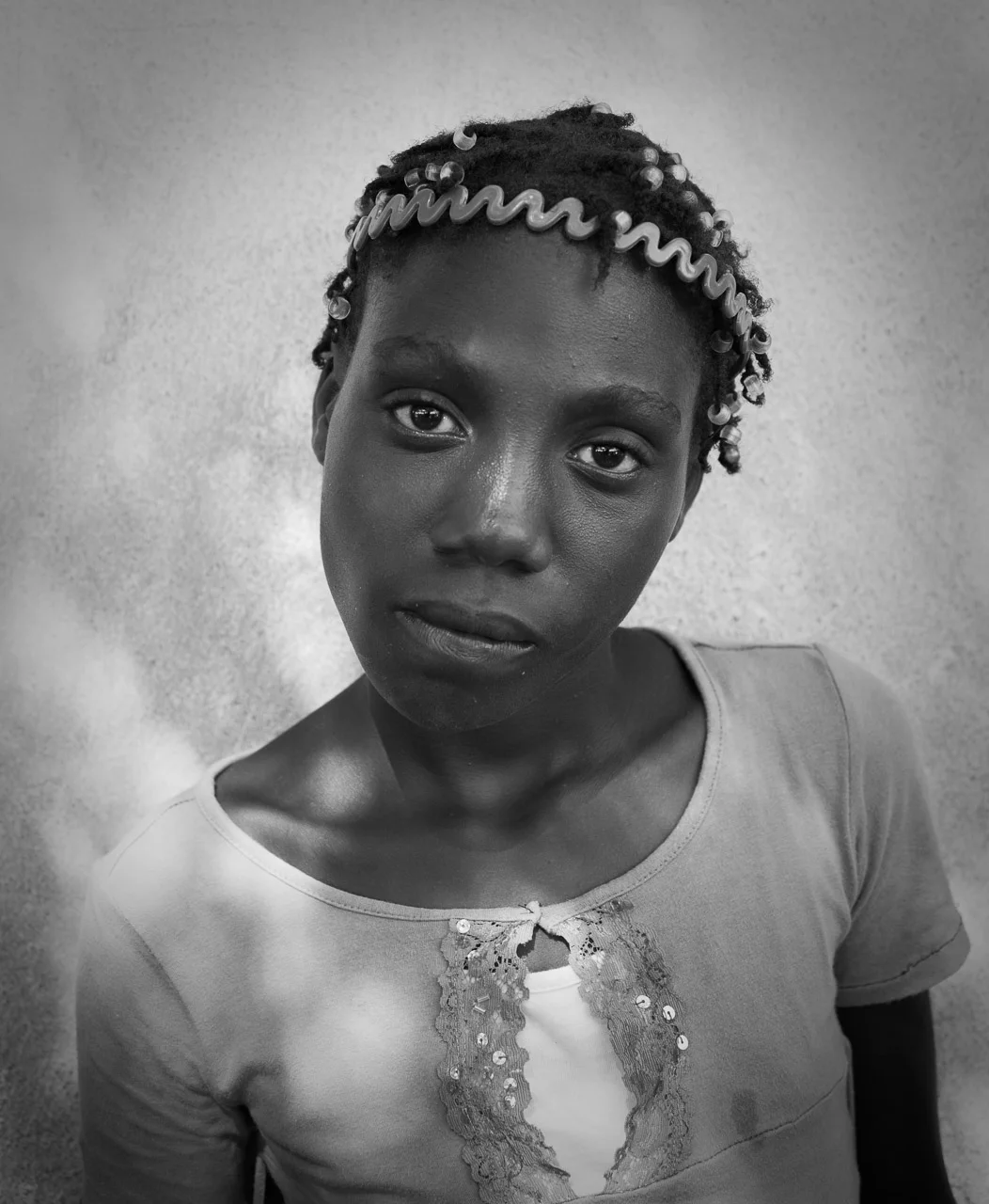 Black and white portrait of a young girl with short curly hair decorated with beads and a spiral headband, wearing a top with lace and sequin embellishments.