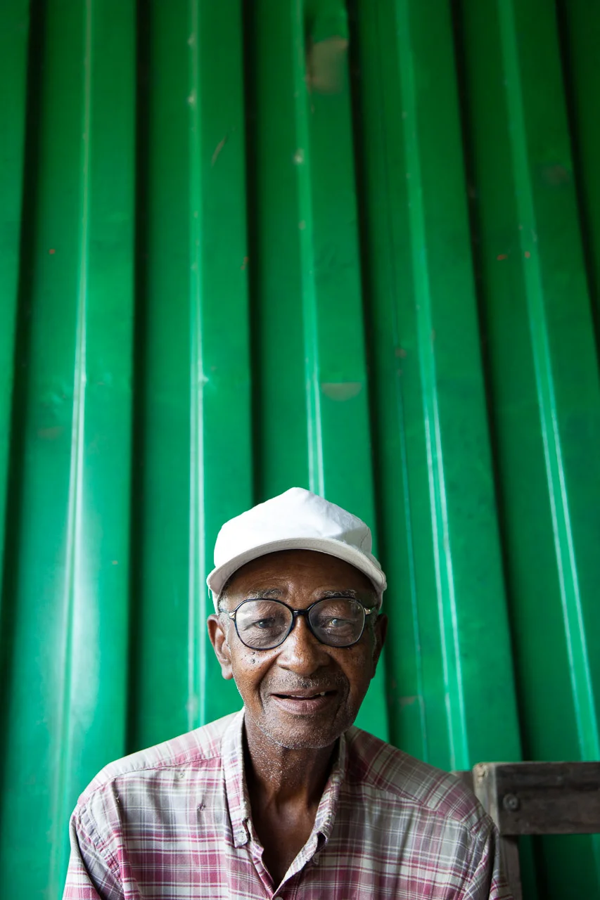 Older man wearing glasses and a white cap, sitting in front of a green corrugated metal wall, smiling.
