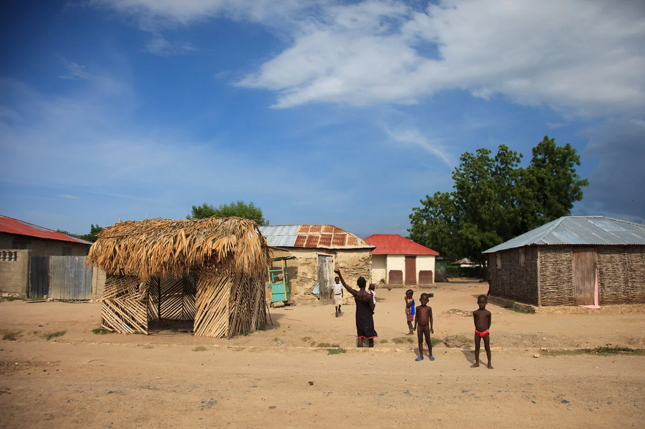 A village scene with several children playing outside near traditional thatched and metal-roofed houses under a blue sky with clouds.