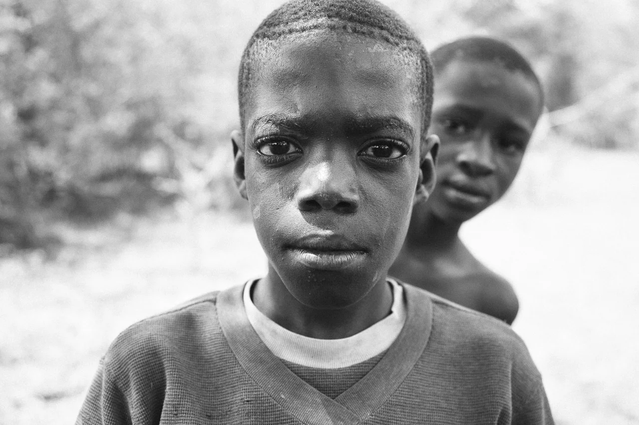 Close-up of two young boys looking at the camera outdoors, one in the foreground and the other slightly behind, in black and white.