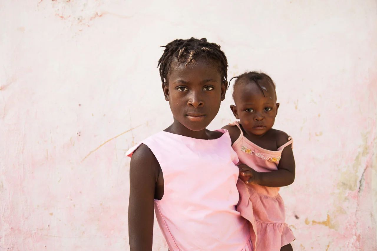 A young girl holding a toddler girl against a pink wall.
