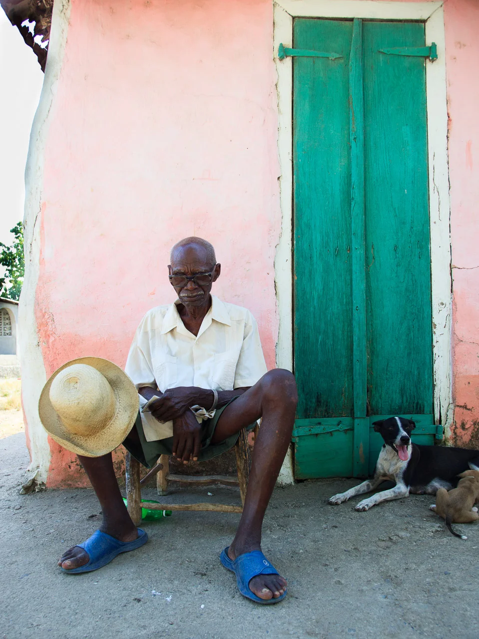 An elderly man with glasses and a white shirt sitting on a small wooden chair outside a pink wall with a green door. He is holding a straw hat and has a dog lying on the ground beside him, with other puppies nearby.