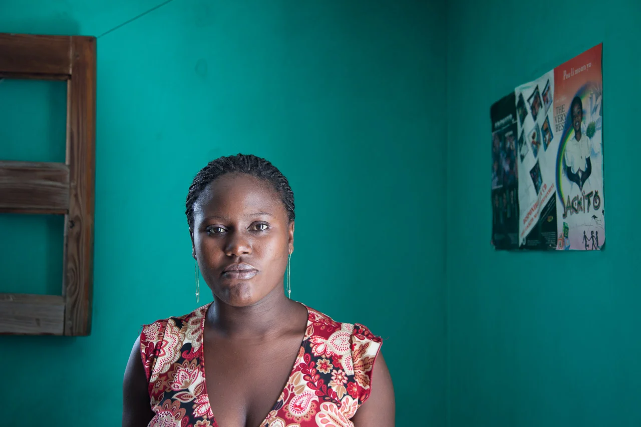 Haitian woman with dark hair and earrings standing in a room with teal walls and a colorful poster on the wall.