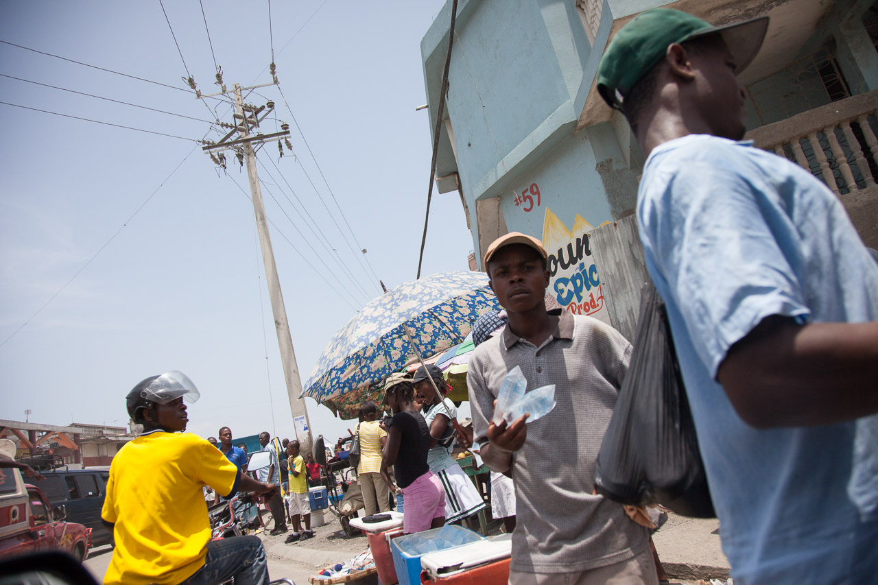 Travel photographer - Haiti scene - People walking and selling goods on a busy street in an urban area, with some individuals carrying umbrellas for shade, and a prominent power pole with wires overhead.