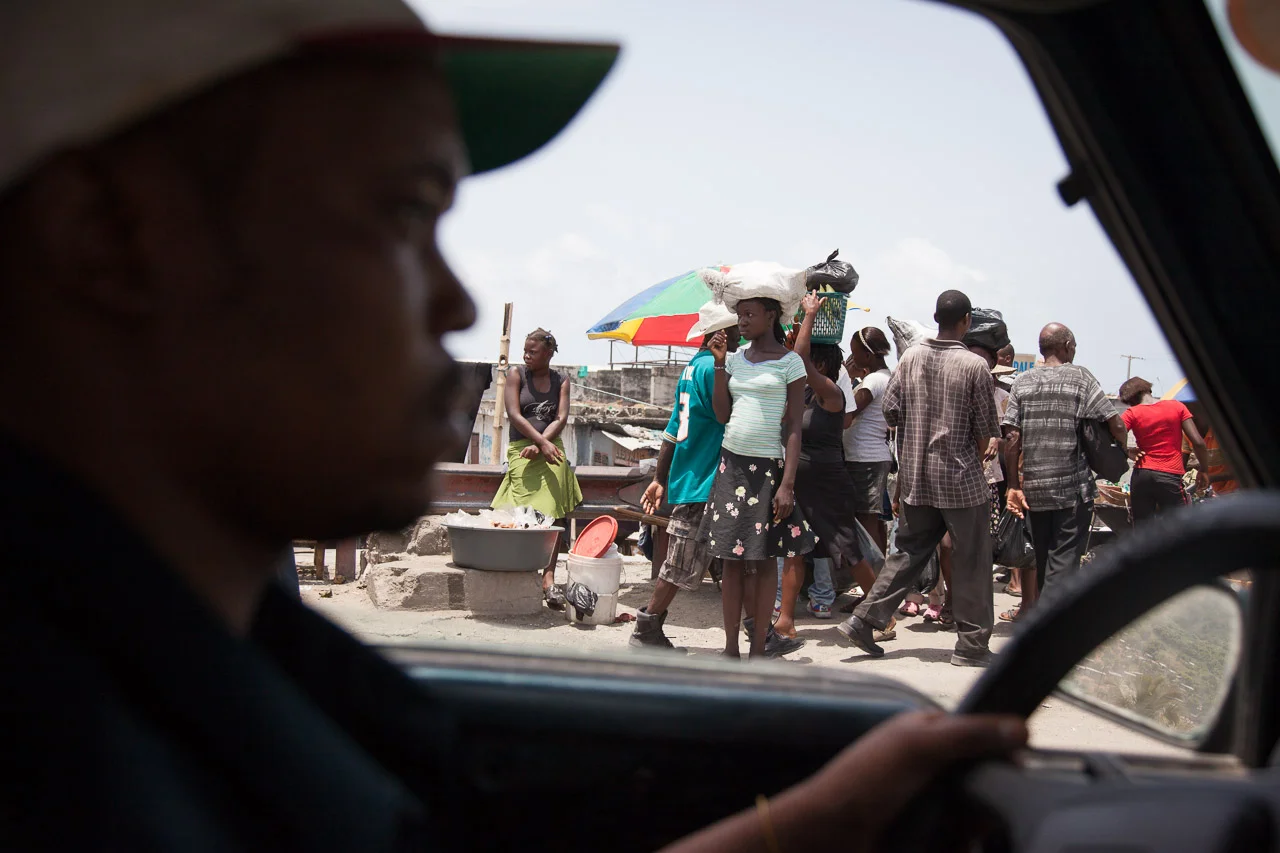 A person sitting in a vehicle looking out at a bustling outdoor market with people walking, carrying goods, and a colorful umbrella in the background.