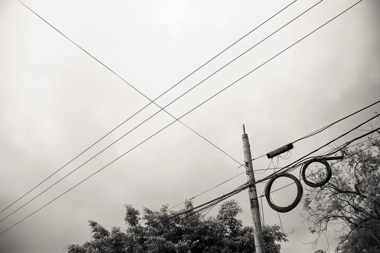 Overhead view of electrical wires and a utility pole with trees in the background under a cloudy sky.