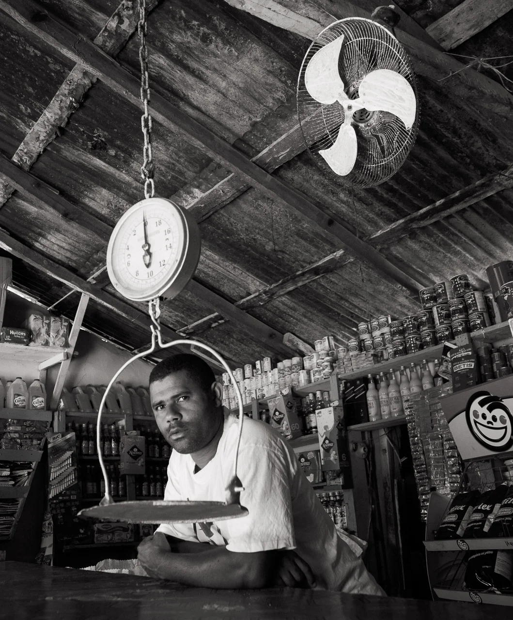 A young man with short hair in a store, leaning on the counter with his arms crossed, surrounded by shelves stocked with canned and bottled goods. A hanging scale and a ceiling fan are visible above him, in a rustic setting.