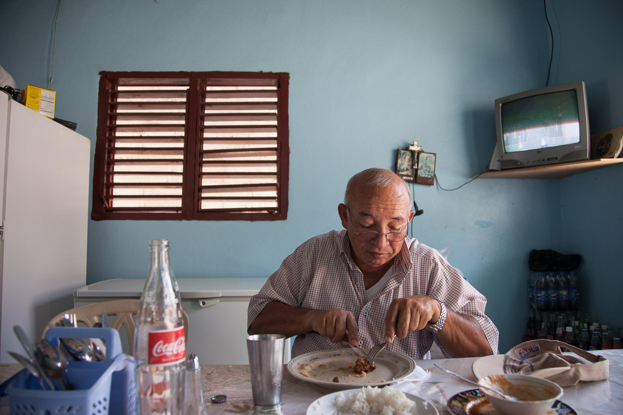 An elderly man is eating food at a dining table in a small room with a blue wall, a red window, a TV, and various items on the table.