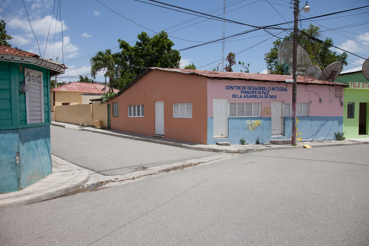 Street corner with pastel-colored buildings, one pink with blue bottom, another green, and a yellow fence, under a blue sky with clouds, with satellite dishes and power lines overhead, in a sunny day.