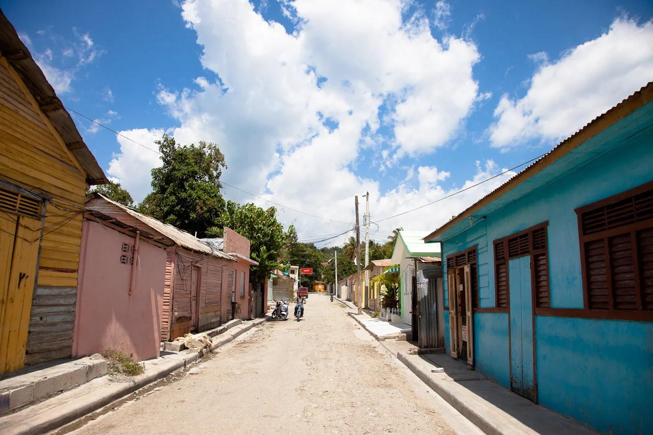 A dirt street lined with colorful small houses under a blue sky with scattered clouds, in a rural or small town setting.