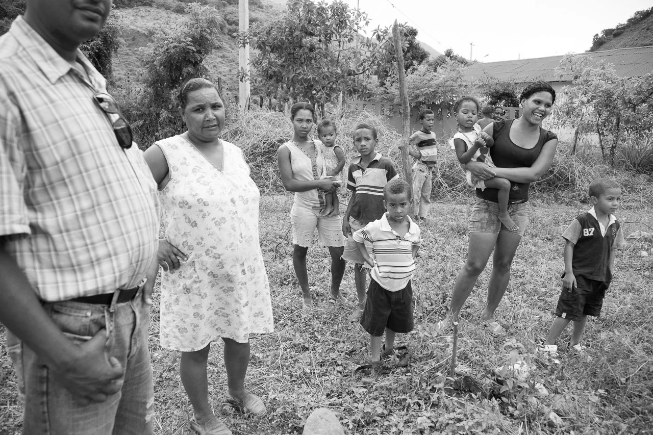 Group of adults and children standing outdoors in a rural area, some looking at the camera and others looking away, with trees and a building in the background.