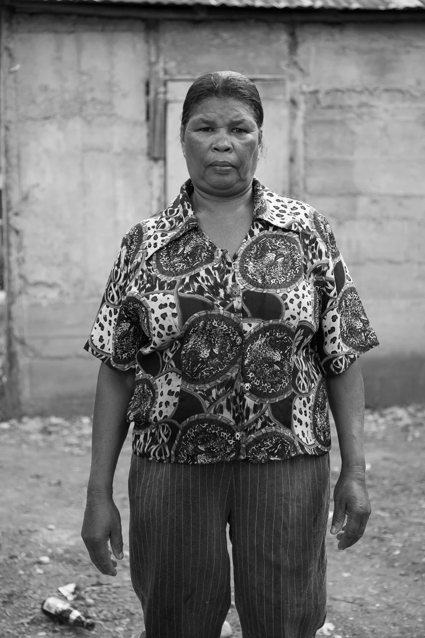 Black and white photo of a woman standing outdoors in front of a wooden wall, wearing a patterned shirt and pinstriped pants, with a serious expression on her face.