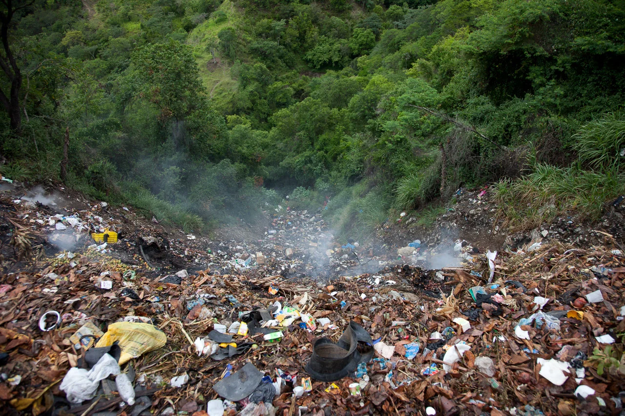 A hill covered with trash and garbage, with smoke rising from the debris in a forested area.