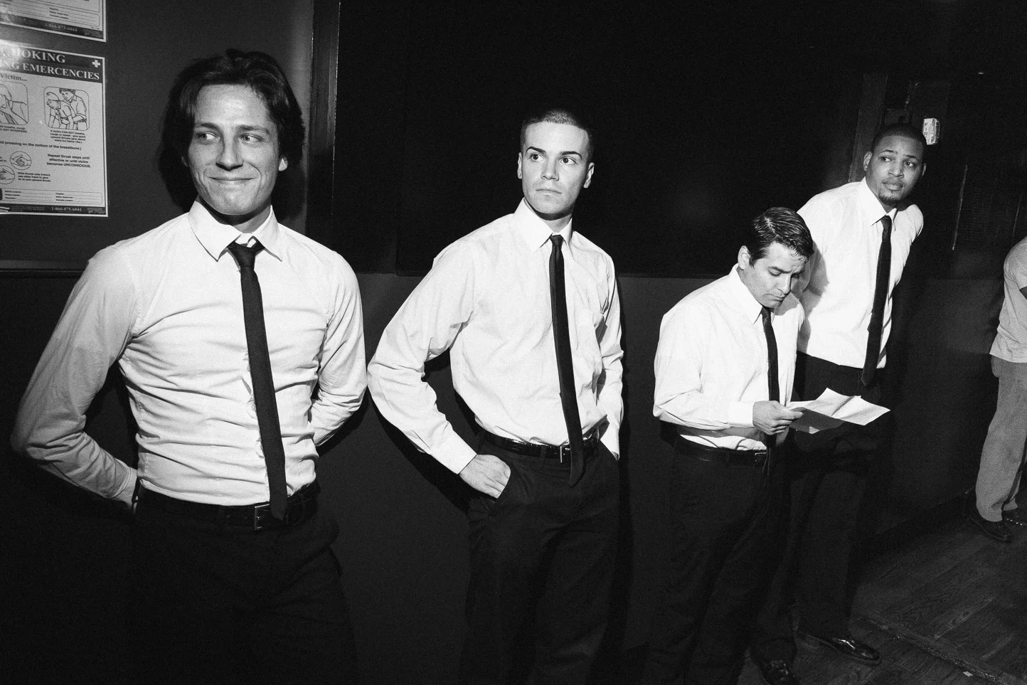 Four men in white shirts and black ties standing in a row, with one reading a paper, in an indoor setting.