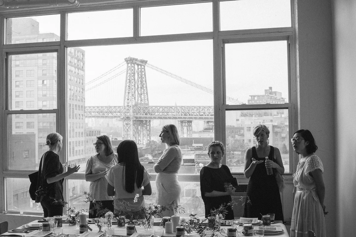 A group of seven women standing near a large window in an indoor setting, with a view of the Brooklyn Bridge and city skyline outside. The women are engaged in conversation, with some holding drinks, and a decorated table visible in the foreground.