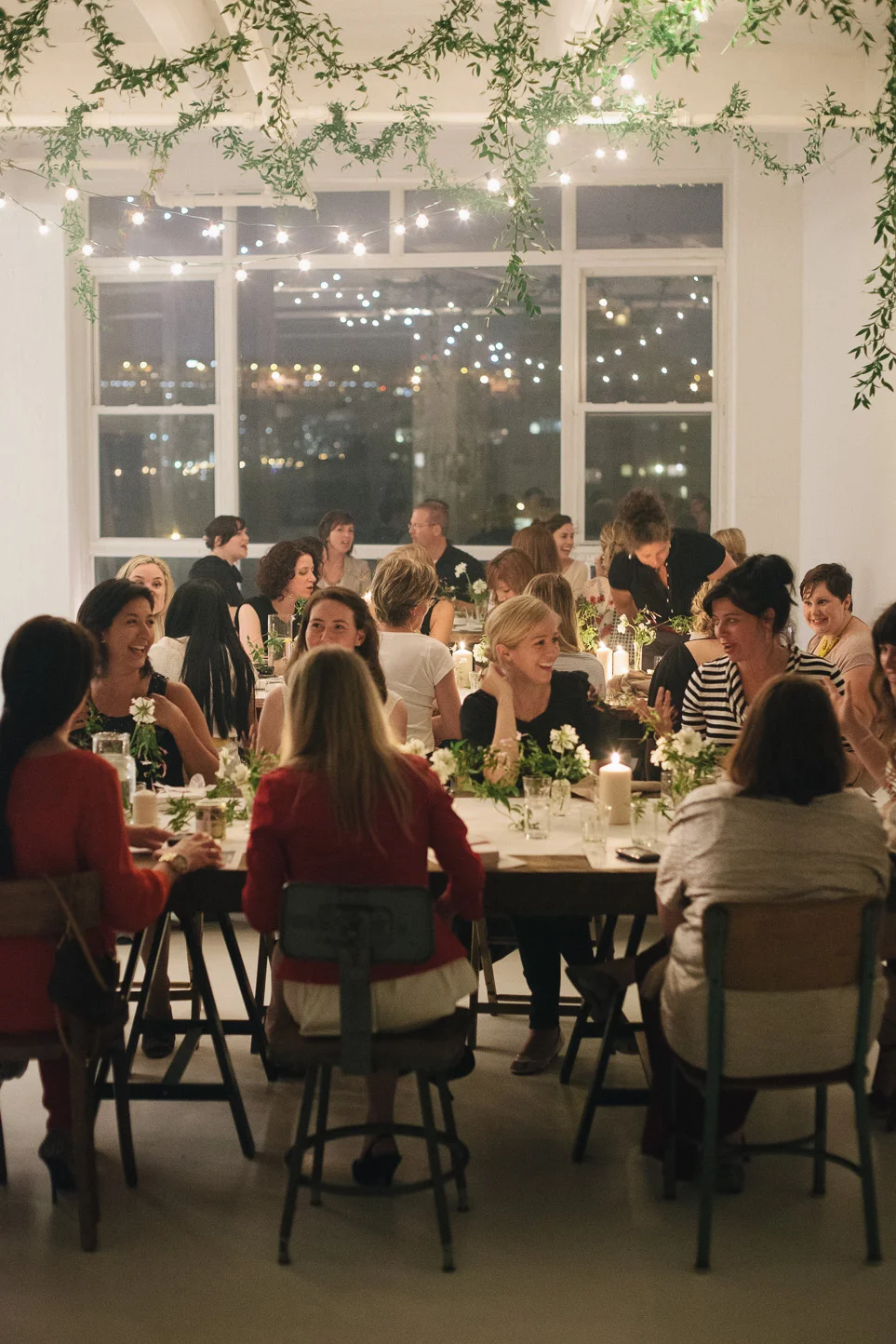 People gathered at a dinner party in a well-lit room with large windows, decorated with hanging green foliage and string lights, with city lights visible outside.