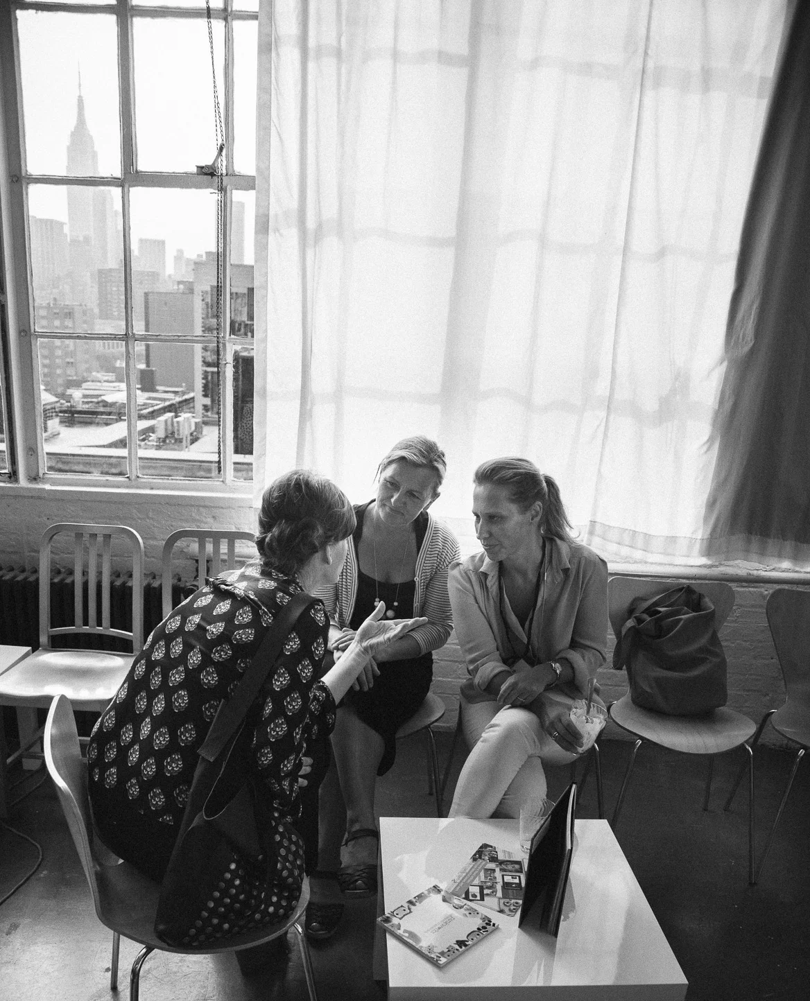Three women engaged in a conversation in a room with large windows showing a cityscape.