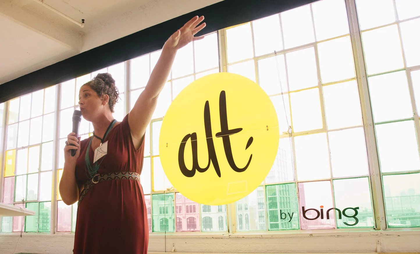 A woman speaking into a microphone during a presentation, standing next to a yellow circle sign with the word 'alt' and the small text 'by bing' in the corner, in front of large multi-colored window panes.