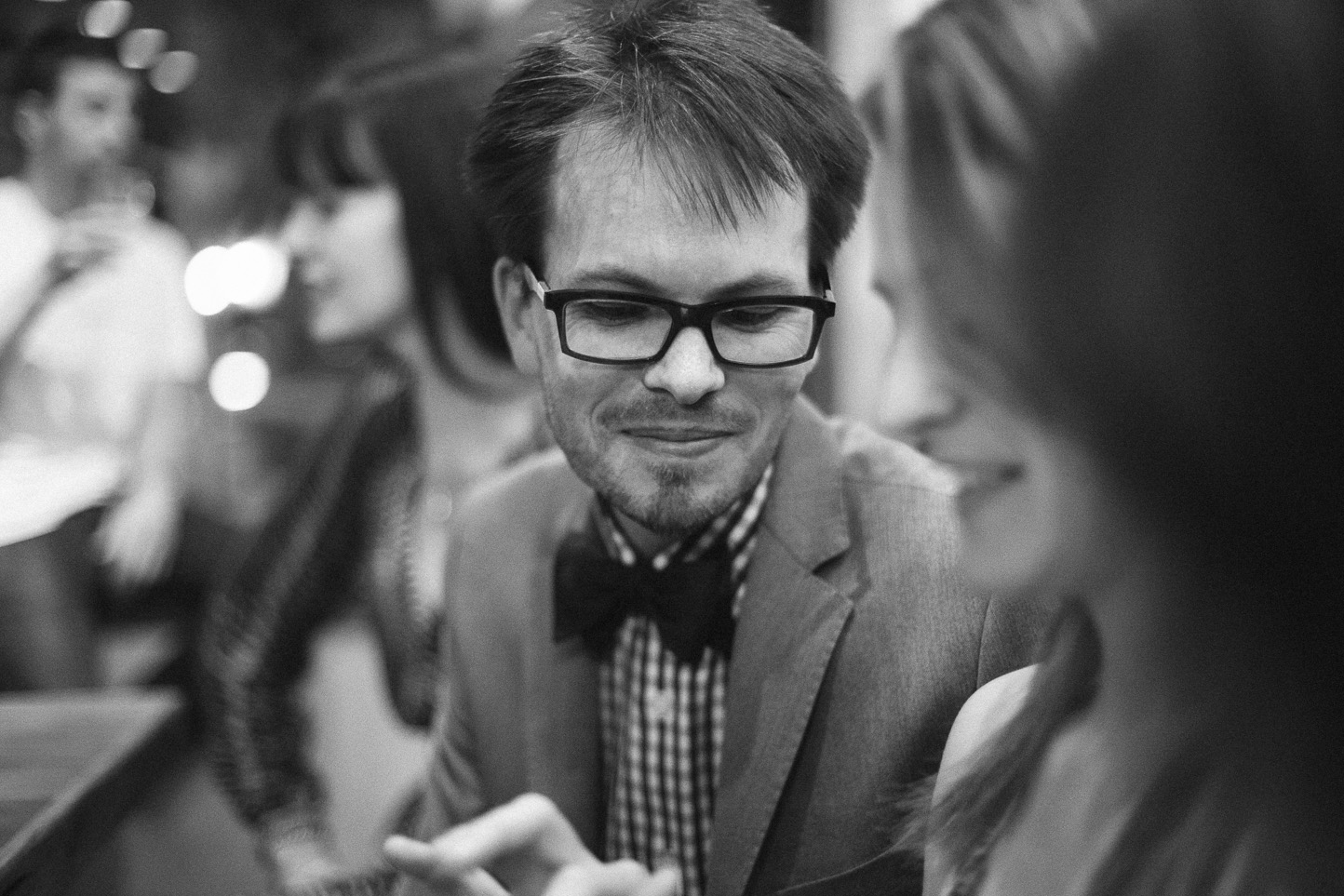 Black and white photograph of a man with glasses and a bow tie smiling and engaged in conversation with a woman, with other people blurred in the background.