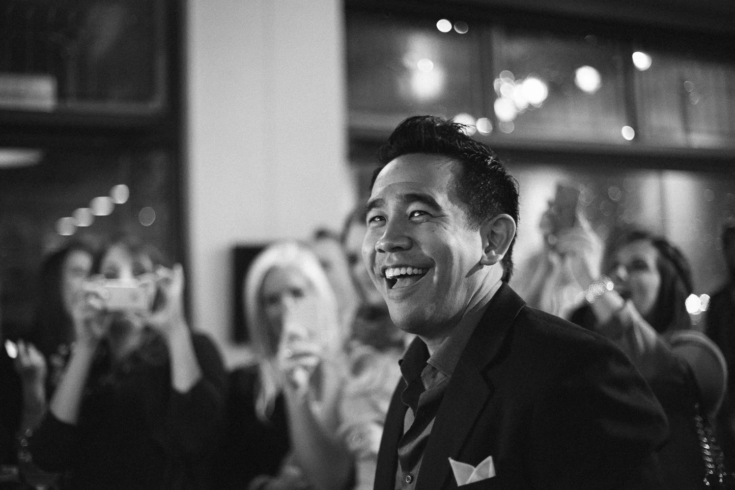 A smiling man in a suit at a social gathering or party, with blurred women in the background taking photos and clapping.