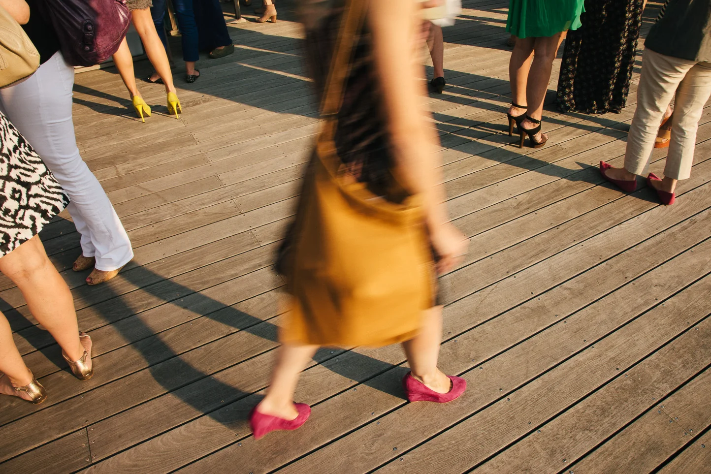 People standing and walking on a wooden deck, mostly focusing on their legs and shoes, with some wearing high heels and others casual footwear.