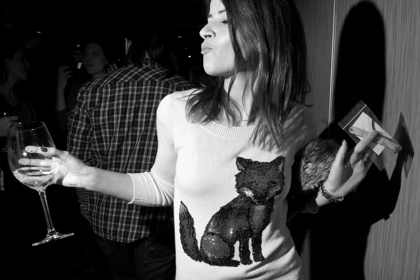 A woman holding a wine glass in one hand and a small bag in the other at a social gathering, wearing a white sweater with a large sequined wolf design on the front.