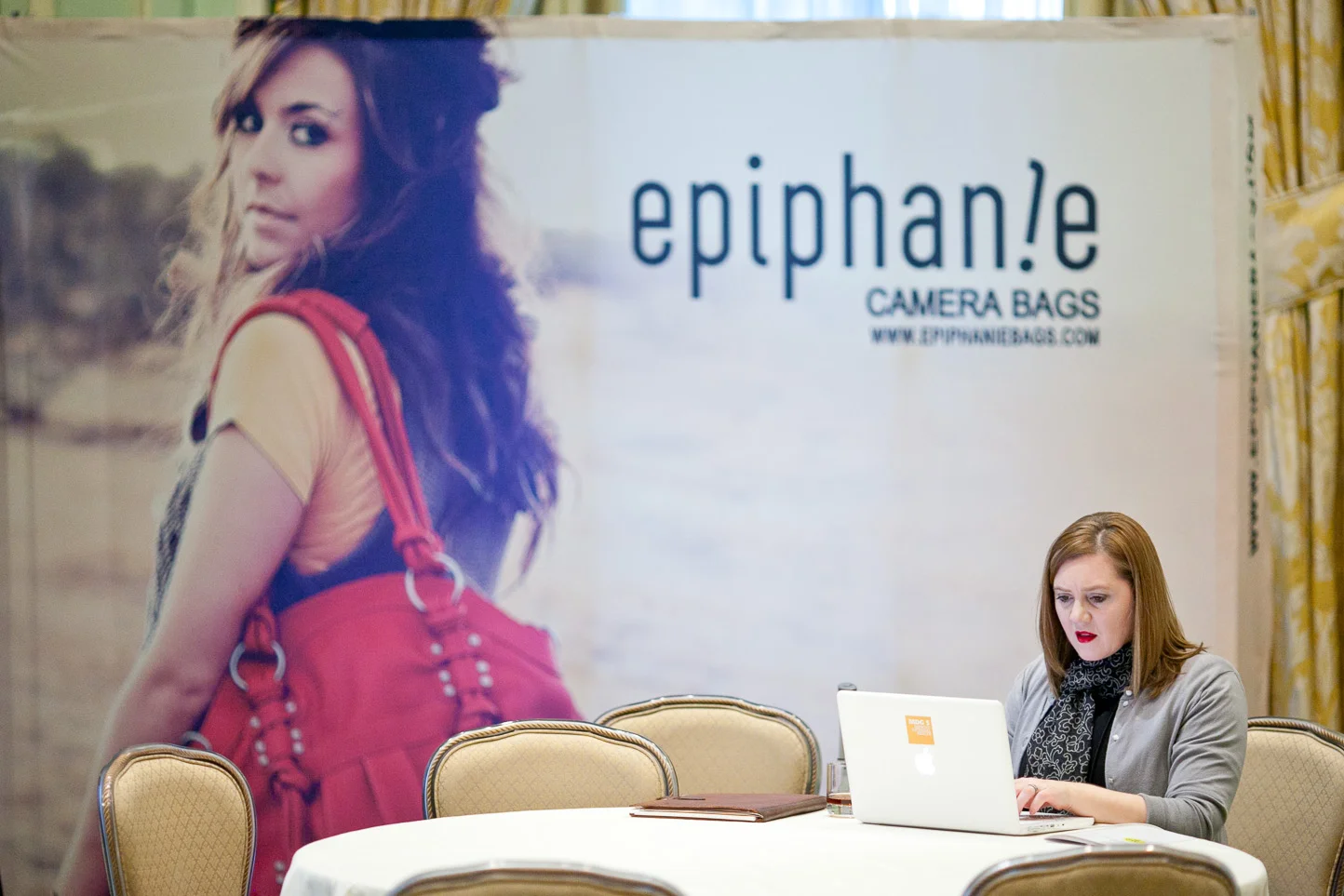 A woman with shoulder-length red hair working on a laptop at a table, with a large banner behind her advertising Epiphan!e camera bags, featuring a woman with long, wavy hair carrying a red camera bag.
