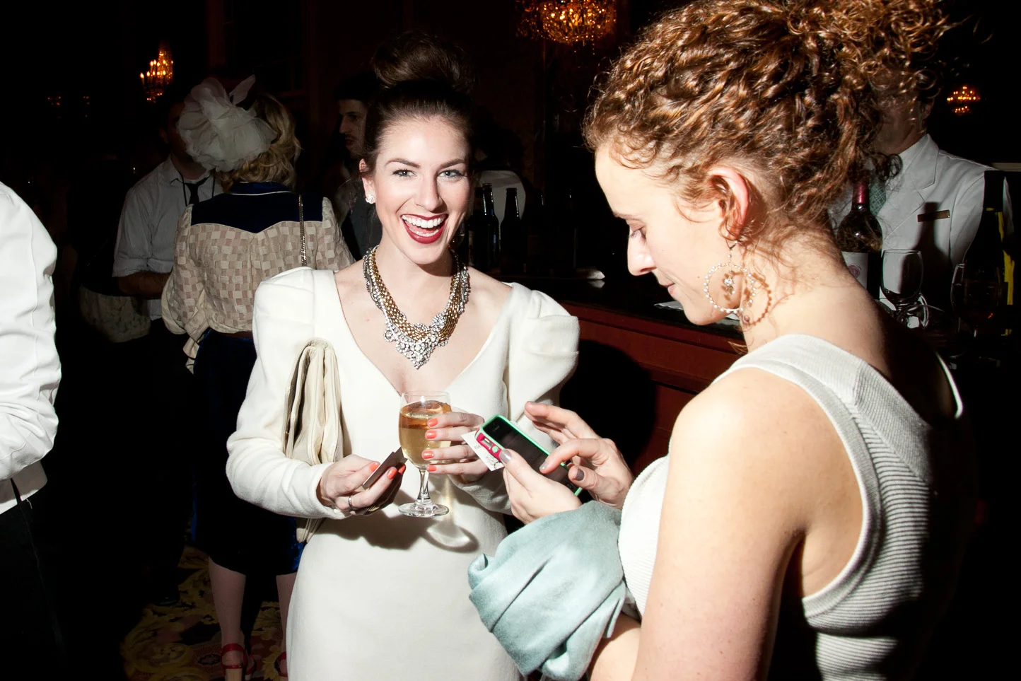 Two women at a party, one in a white dress with jewelry holding a glass of wine, smiling, and another in a sleeveless top looking at her phone.