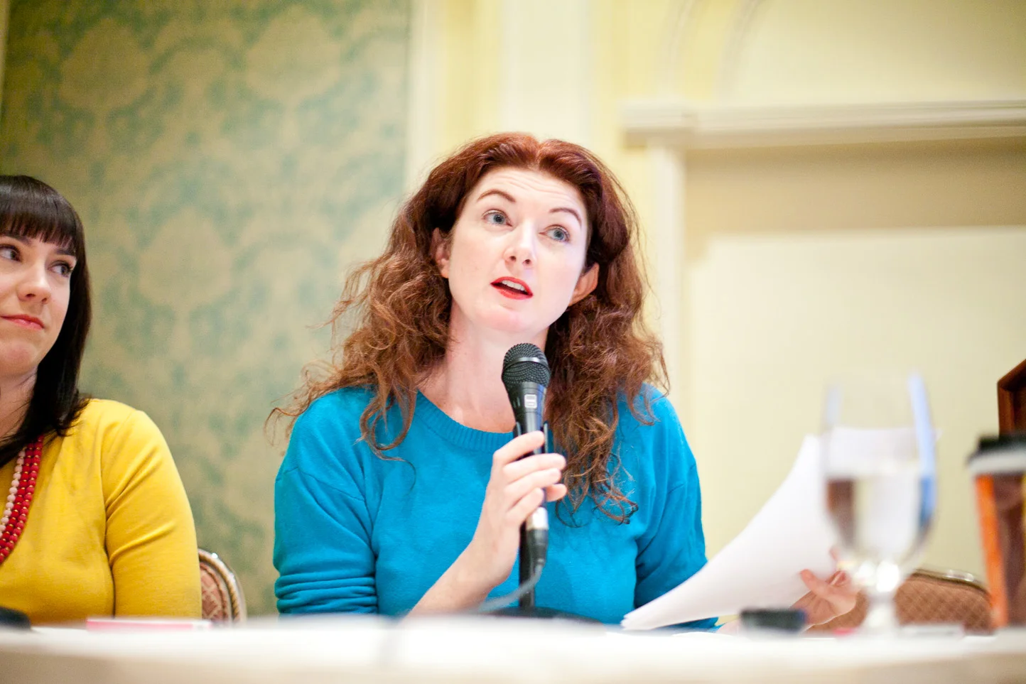 Woman with red hair in a blue shirt speaking into a microphone during a panel or meeting.