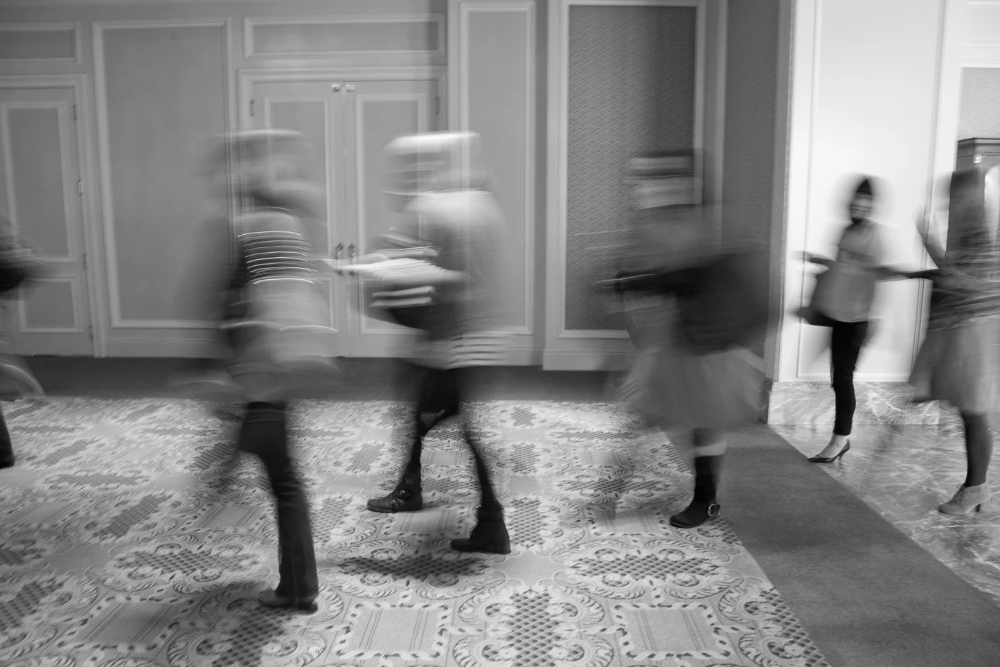 Blurry black and white photo of people walking in a hallway, some holding papers or devices.