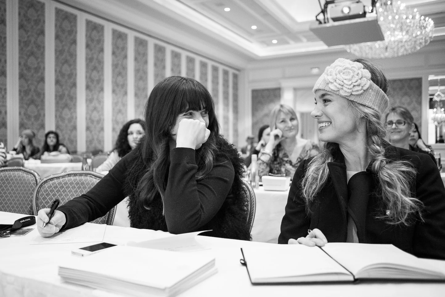 Two women at a conference table smiling and laughing, with others in the background, in a large decorated room with chandeliers.