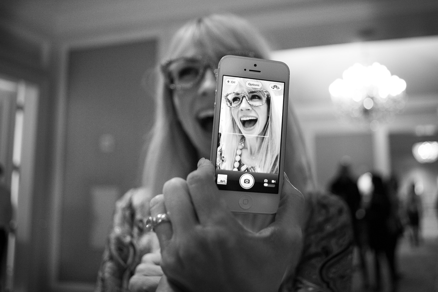 A woman taking a photo with an iPhone shows her smiling and wearing glasses, with a chandelier in the background.