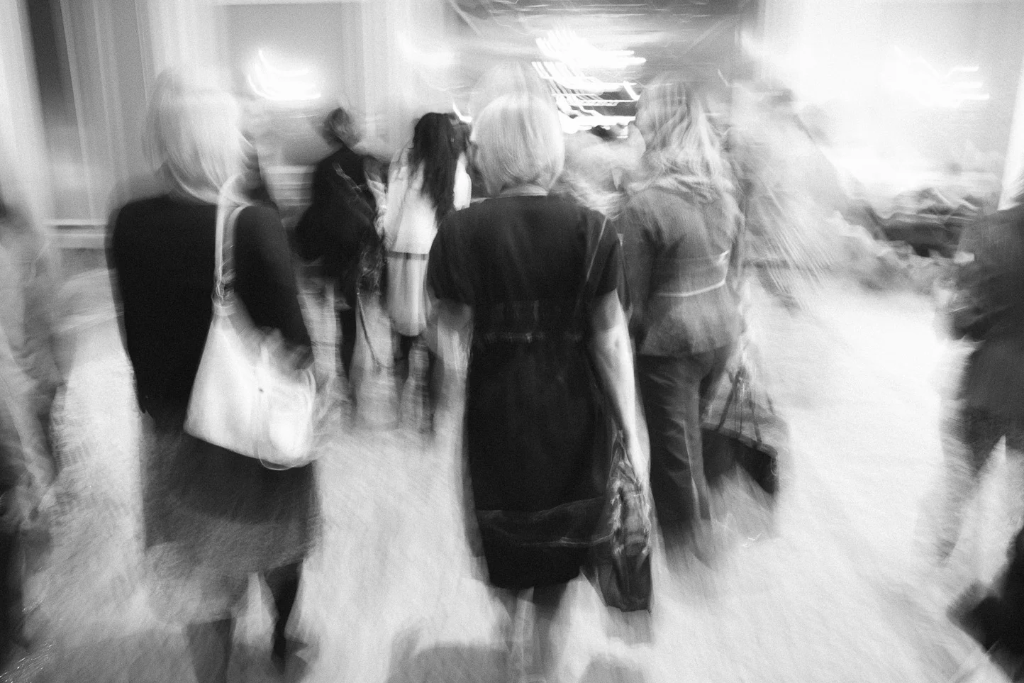 Blurred black and white photo of a group of women walking in a lobby or corridor, carrying bags.