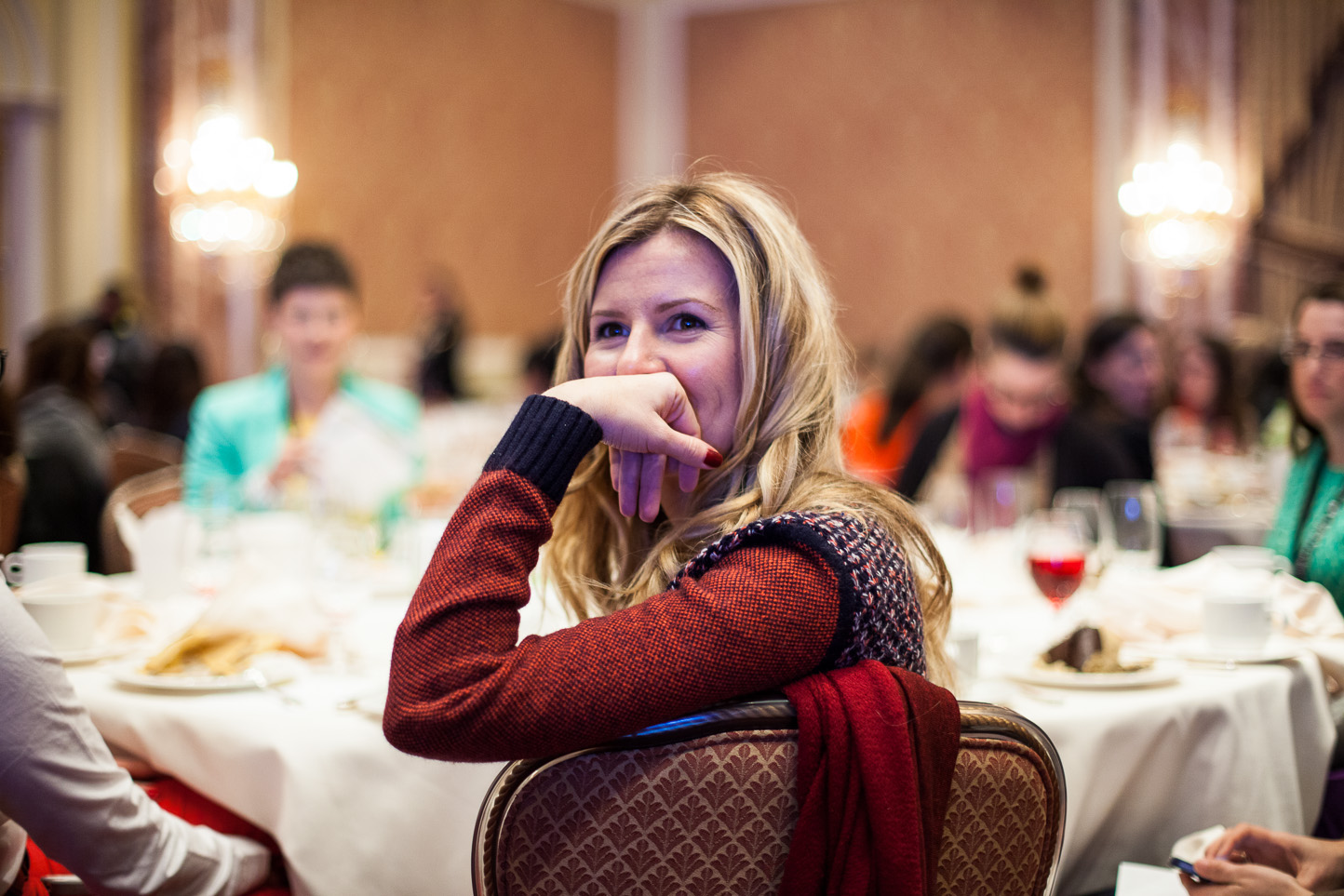 A blonde woman with a red sweater covering her mouth and smiling, sitting at a round banquet table during a social event, with other people in the background.