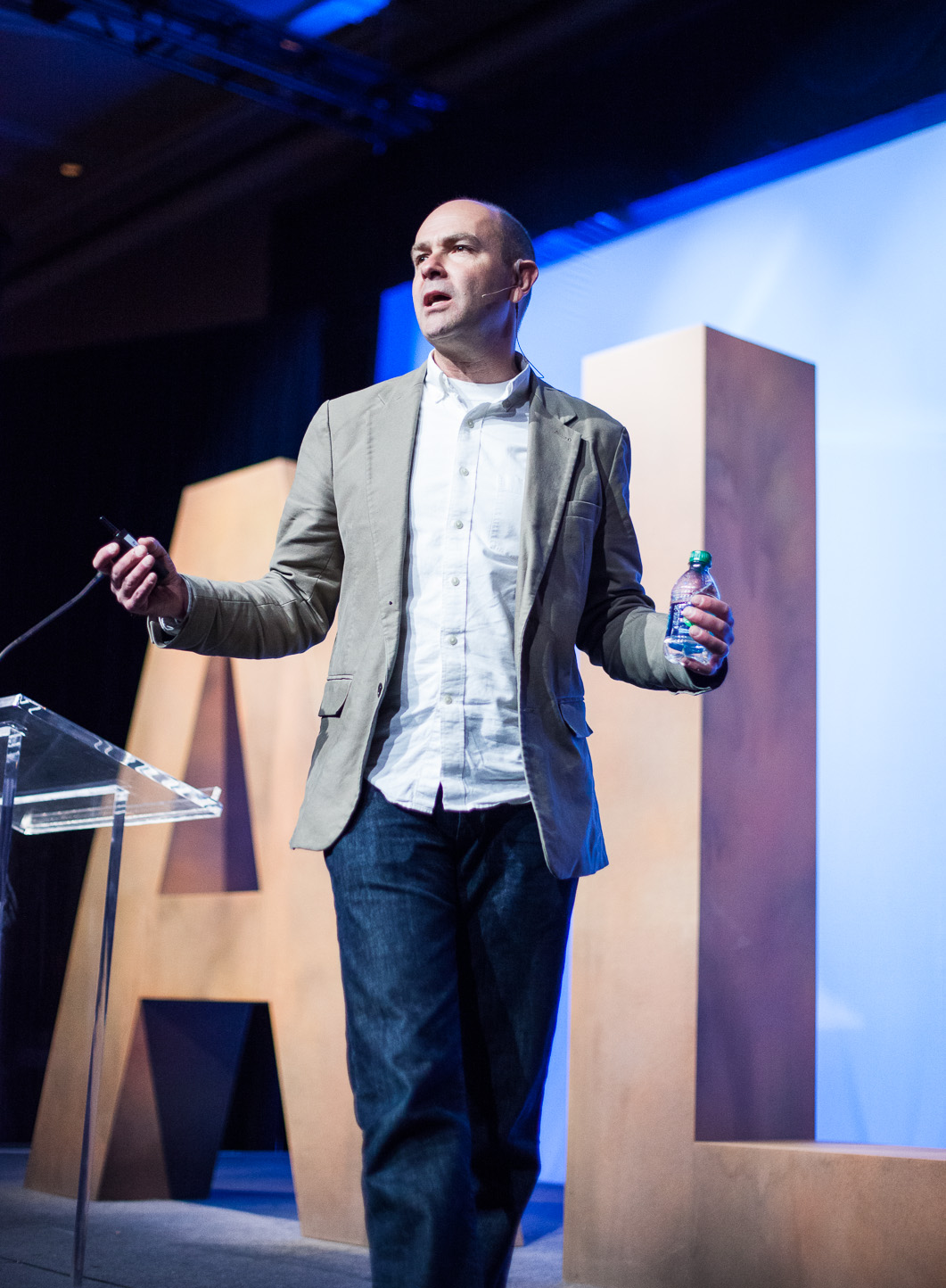 A man giving a presentation on stage, holding a water bottle in one hand and a remote in the other, with large wooden letters in the background.