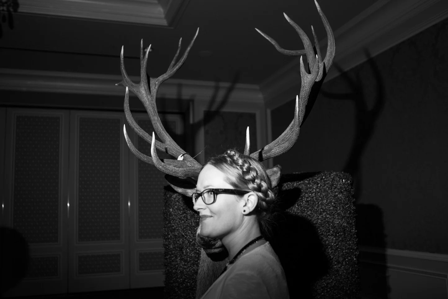 A woman with glasses and braided hair wearing a deer antler headpiece, sitting in profile against a textured backdrop in a dimly lit room.