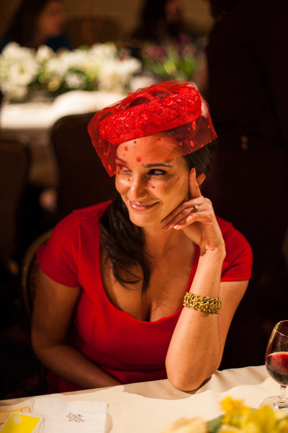 A woman in a red dress with dark hair, wearing a red veil with polka dots and a gold bracelet, sitting at a table during a festive event, smiling with her hand near her face.