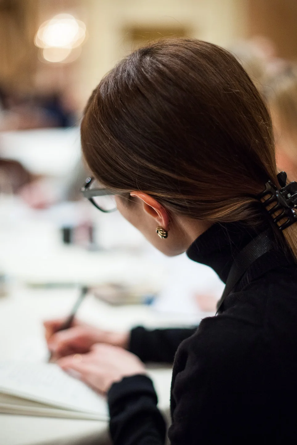 A woman with brown hair tied back, wearing glasses, earrings, and a black top, writing in a notebook at a table in a busy indoor setting.