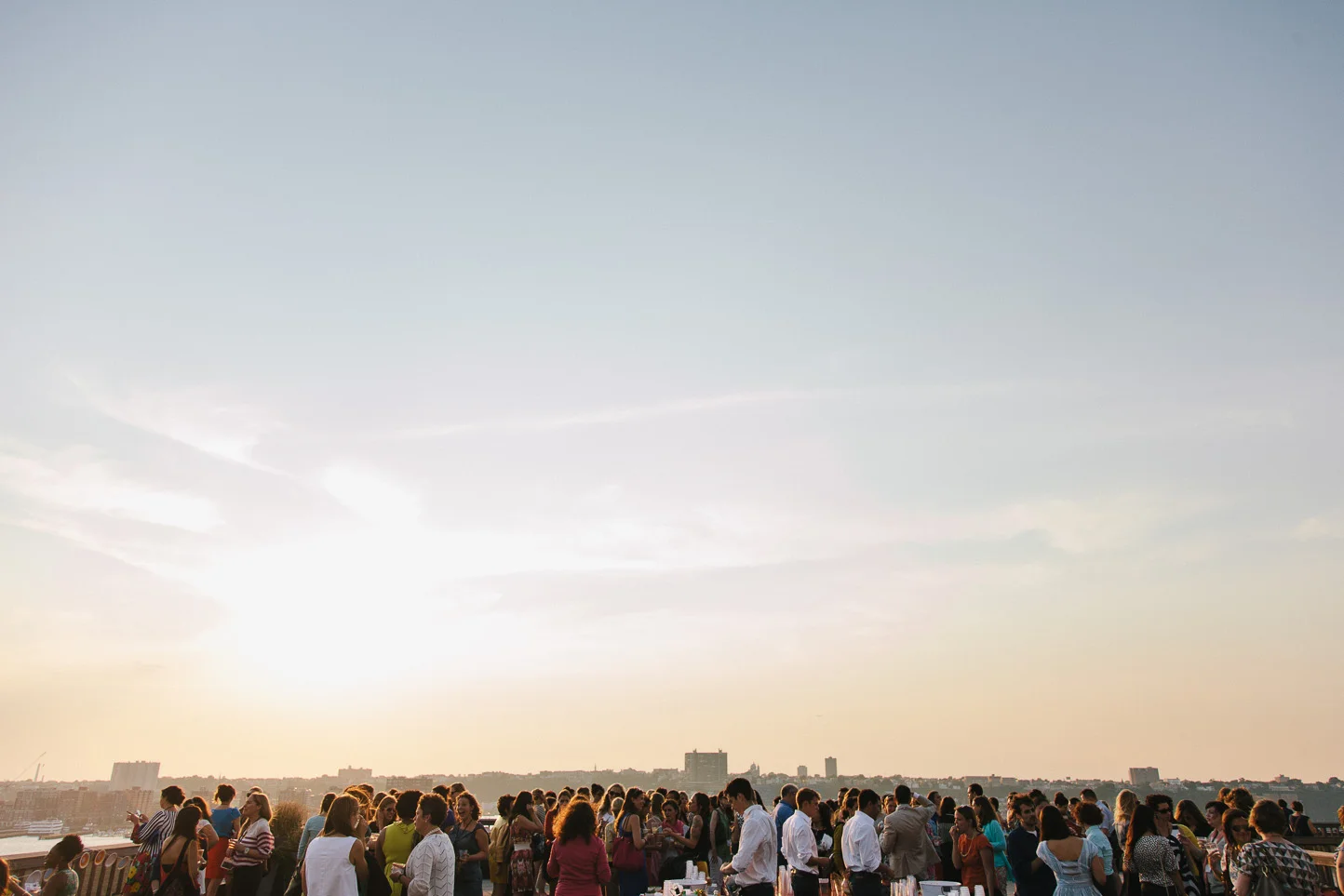 Crowd of people gathered on a rooftop during sunset, with city skyline in the background.