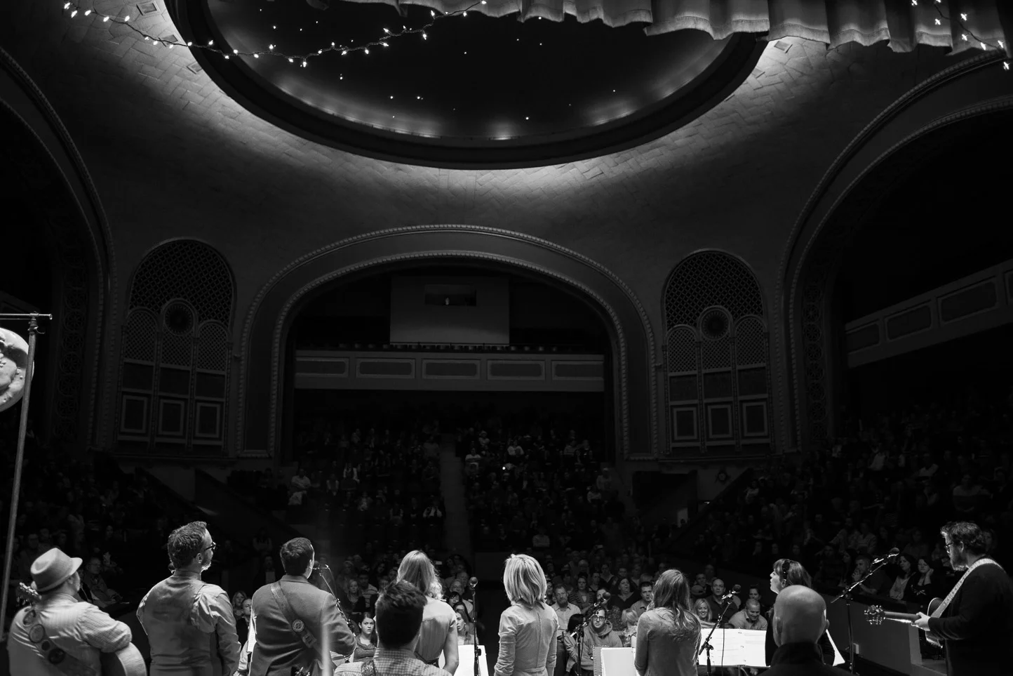 Musicians performing on stage inside a theater with an audience in attendance, viewed from the stage.