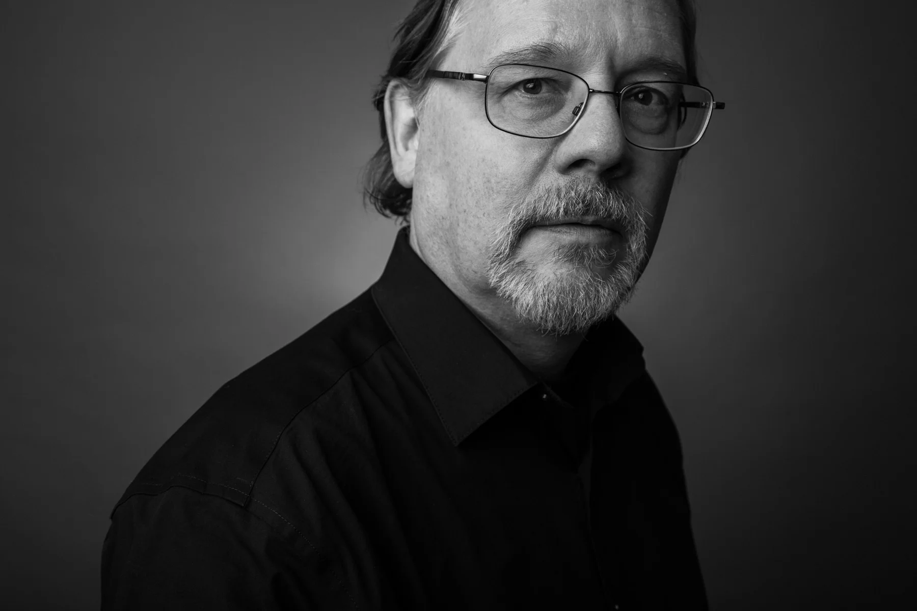 Black and white portrait of a middle-aged man with glasses, a beard, and mustache, wearing a black shirt, against a dark background.