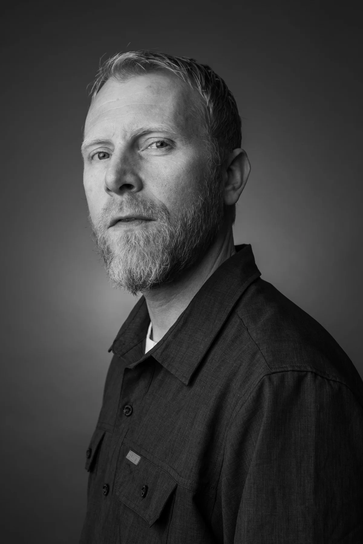 Black and white portrait of a bearded man with short hair, wearing a button-up shirt, looking at the camera against a plain background.