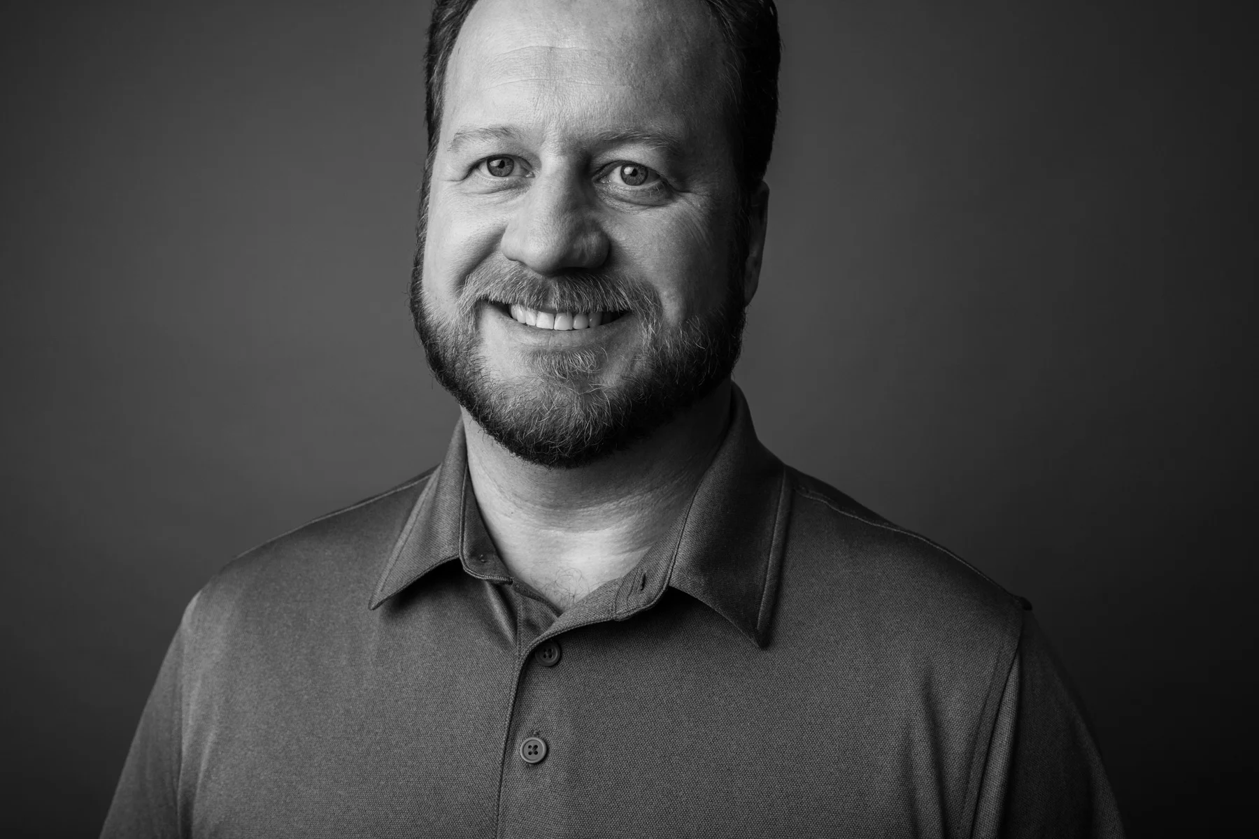 Black and white portrait of a smiling man with a beard, wearing a collared shirt, facing the camera against a plain background.