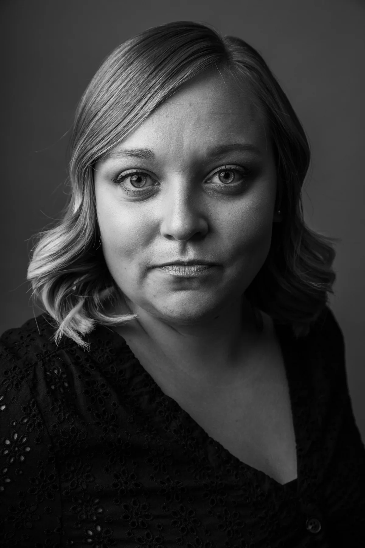 Black and white portrait of a woman with wavy hair, wearing a black eyelet lace top, looking directly at the camera.