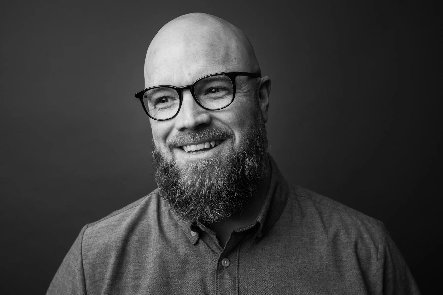 Black and white portrait of a smiling bald man with a beard wearing glasses and a button-down shirt against a dark background.